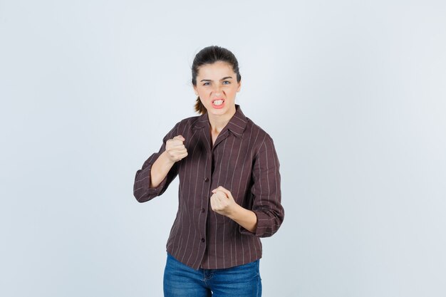 femme debout dans la pose de combat, serrant les dents en chemise, jeans et semblant agressive, vue de face.