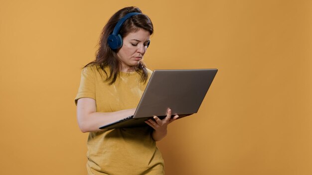 Femme décontractée portant des écouteurs sans fil tenant un ordinateur portable et tapant sur le clavier en regardant l'écran debout en studio. Personne détendue écoutant de la musique sur des écouteurs à l'aide d'un ordinateur portable.
