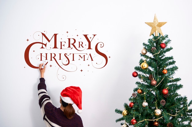 Femme décorant un arbre de Noël et écrivant un texte de joyeux Noël sur le mur le jour de Noël