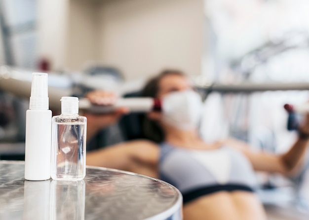 Femme défocalisée à la salle de sport à l'aide d'équipement avec masque médical et bouteille de désinfectant pour les mains