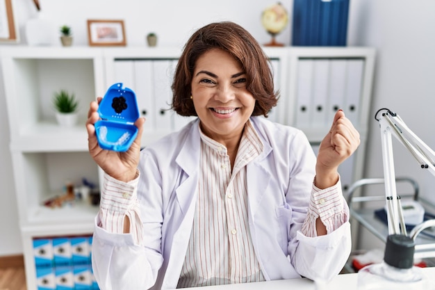 Femme dentiste hispanique d'âge moyen travaillant à la clinique dentaire tenant un aligneur invisible criant fièrement célébrant la victoire et le succès très excitée avec les bras levés