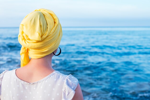 Femme de derrière avec un foulard jaune couvrant sa tête sans cheveux en contemplant l'horizon de la mer