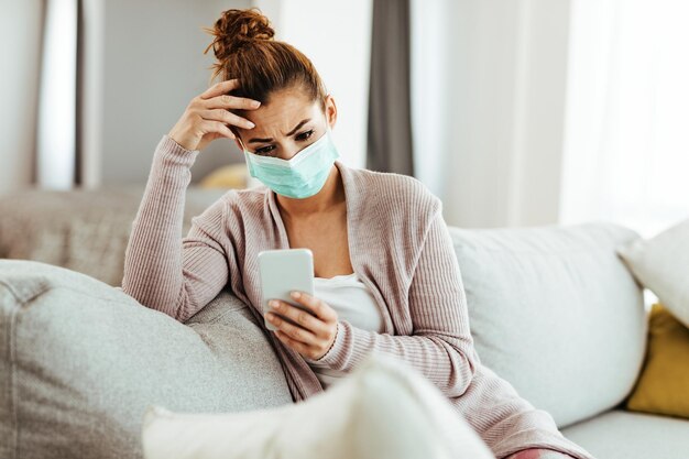 Femme désemparée portant un masque de protection assise sur le canapé et envoyant des SMS sur son téléphone portable à la maison