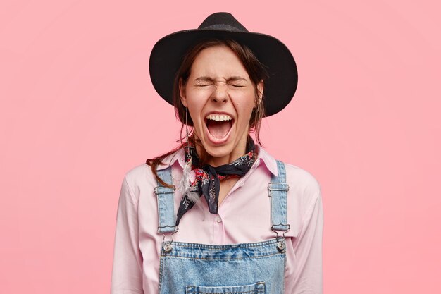 Une femme désespérée, propriétaire d'un ranch, hurle de désespoir, a des problèmes financiers, ne peut pas tout gérer seule, porte des vêtements de campagne, se tient contre un mur rose