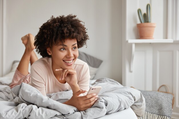 Femme détendue à la peau sombre avec une coiffure afro, garde les jambes levées, habillée en pyjama, tient un téléphone cellulaire moderne, a une expression rêveuse, navigue sur Internet, envoie des messages texte, profite d'une atmosphère calme