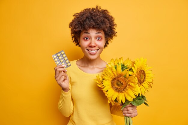 une femme détient des médicaments et un bouquet de tournesols souffre de rhinite allergique et de rougeur des yeux vêtue avec désinvolture isolée sur un jaune vif