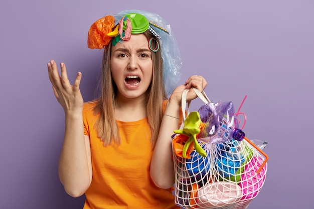 Une femme en difficulté caucasienne fait des gestes avec colère, essaie de nettoyer la planète des déchets en plastique, crie désespérément, tient un sac plein de déchets, porte un t-shirt orange, se tient contre un mur violet.