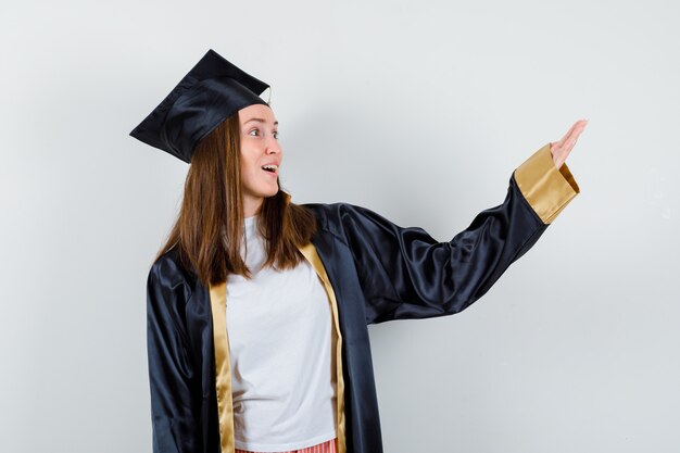 Femme diplômée faisant semblant de montrer quelque chose en tenue académique et à la vue étonnée, de face.