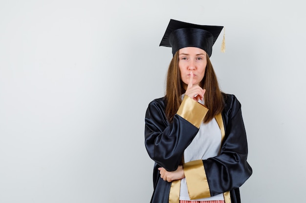 Femme diplômée montrant le geste de silence en uniforme, vêtements décontractés et à la vue sensible, de face.