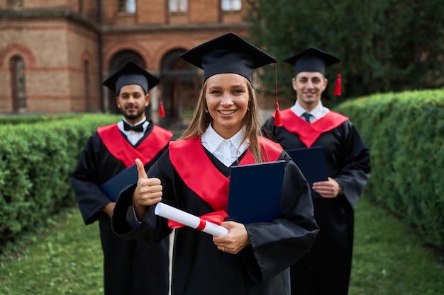 Une femme diplômée montre comme avec ses amis en robes de graduation titulaires d'un diplôme et souriant à la caméra.