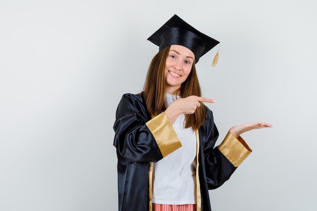 Femme diplômée pointant de côté, faisant semblant de montrer quelque chose dans des vêtements décontractés, uniforme et regardant gai, vue de face.