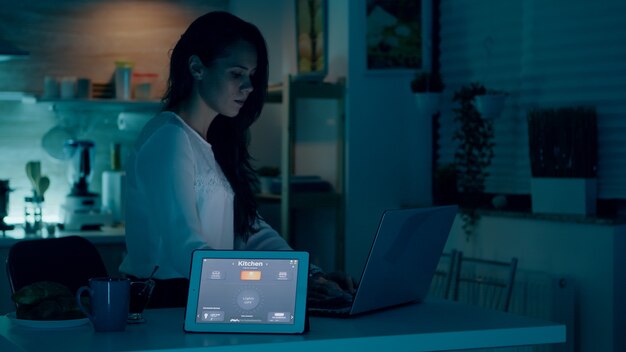 Femme à distance travaillant dans une maison moderne donnant une commande vocale à une tablette avec une application de maison intelligente et des lumières allumées