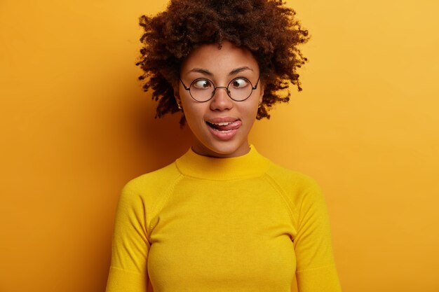 Femme drôle enfantine aux cheveux afro sort la langue, croise les yeux, devient folle et folle, fait la grimace, porte des lunettes rondes et un pull décontracté, pose contre le mur jaune, a une humeur ludique