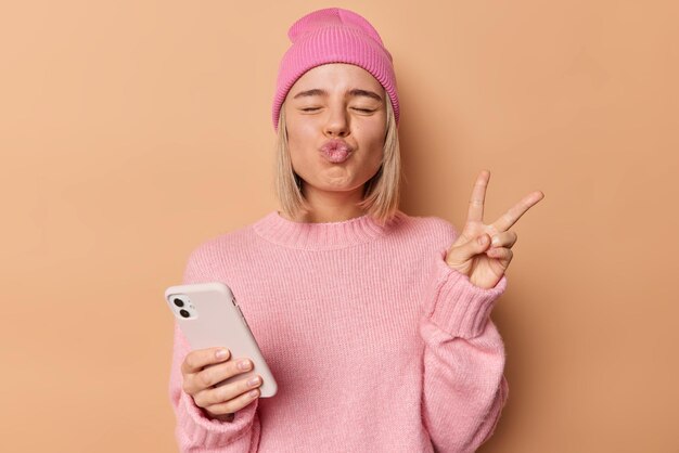 Une femme drôle garde les yeux fermés, les lèvres pliées, fait un geste de paix, tient un téléphone portable, aime la communication en ligne, montre le signe de la victoire vêtu d'un pull décontracté et d'un chapeau isolé sur fond beige.