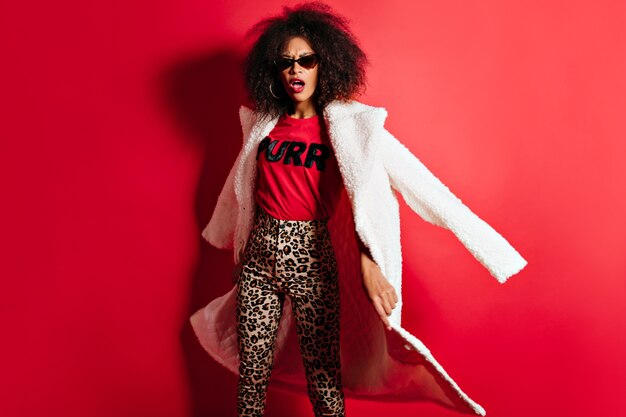 Femme drôle en lunettes de soleil posant sur un mur rouge avec la bouche ouverte