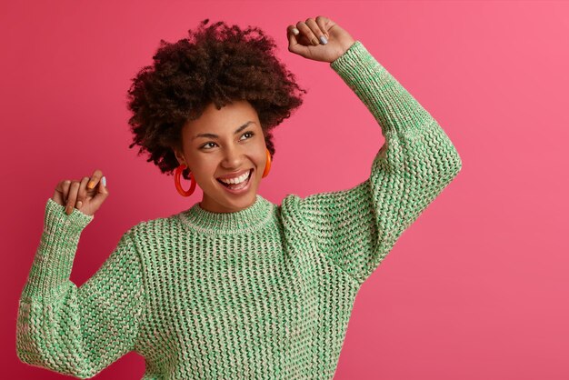 Une femme du millénaire à la peau sombre et insouciante énergique fait danser la victoire, célèbre quelque chose, se sent très heureuse, garde les mains levées, vêtue d'un pull décontracté, a un visage détendu, un large sourire à pleines dents