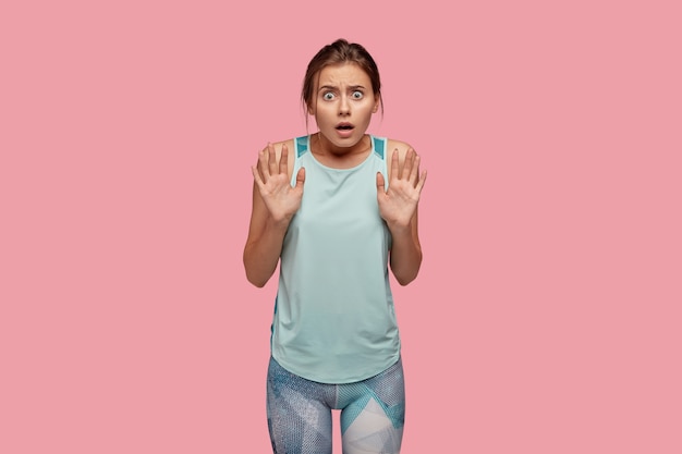 Femme effrayée aux yeux pleins de peur, garde les mains devant, montre un geste protecteur, porte une veste et des leggings décontractés, des modèles sur un mur rose. Une dame stupéfaite et débordée excitée pose à l'intérieur