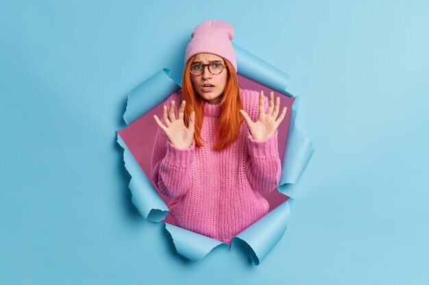 Une femme effrayée et effrayée lève les mains dans un geste défensif voit quelque chose d'horrible porte un chapeau rose et un pull traverse le papier exprime des émotions négatives.