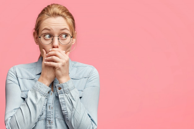 Femme effrayée essaie de se taire, garde les mains sur la bouche, regarde avec une expression inquiète, vêtue d'une veste en jean décontractée, isolée sur rose