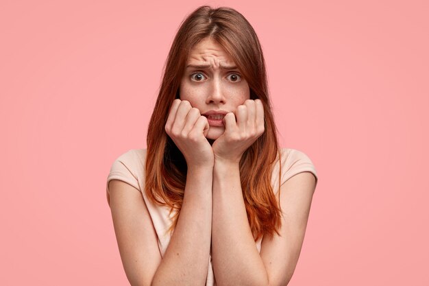 Femme effrayée avec des taches de rousseur sur le visage, regarde avec une expression terrifiée porte un t-shirt rayé décontracté, isolé sur fond rose.