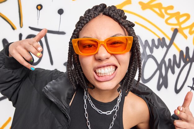 Une femme effrontée avec des dreadlocks serre les dents fait que votre geste se sent cool appartient au gang de rue porte des lunettes de soleil orange à la mode veste à la mode pose contre le mur de graffitis