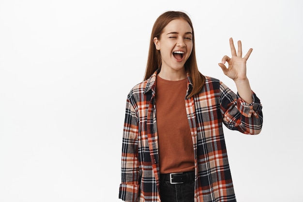 Une femme effrontée souriante fait un clin d'œil et montre un signe OK, assure que tout va bien, sous contrôle, faisant allusion à des remises intéressantes, debout contre un mur blanc.