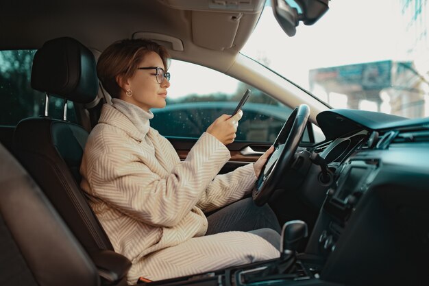 femme élégante assise dans une voiture vêtue d'un manteau d'hiver et de lunettes à l'aide d'un smartphone