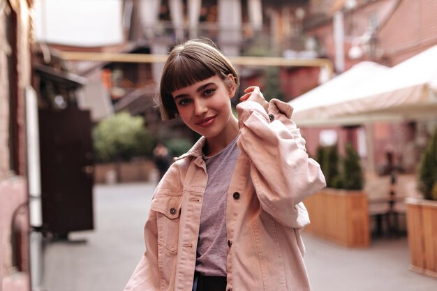 Femme élégante aux cheveux courts en veste en jean souriant dans la ville Femme aux cheveux brune en tenue moderne beige posant en plein air