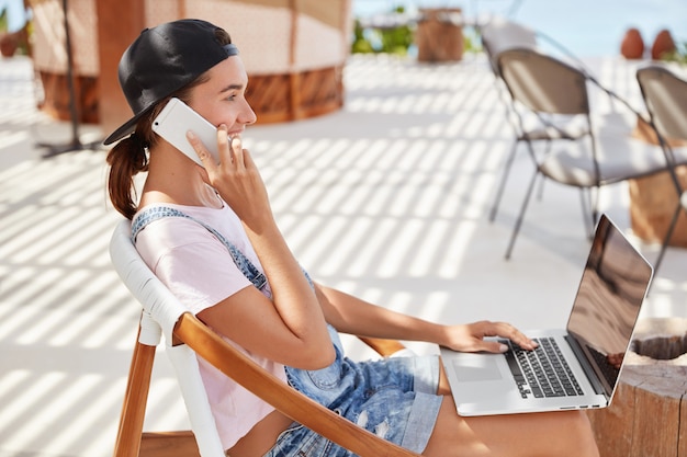 Femme élégante en casquette noire, t-shirt blanc décontracté, messages dans le chat en ligne sur ordinateur portable, parle avec le meilleur ami sur téléphone intelligent