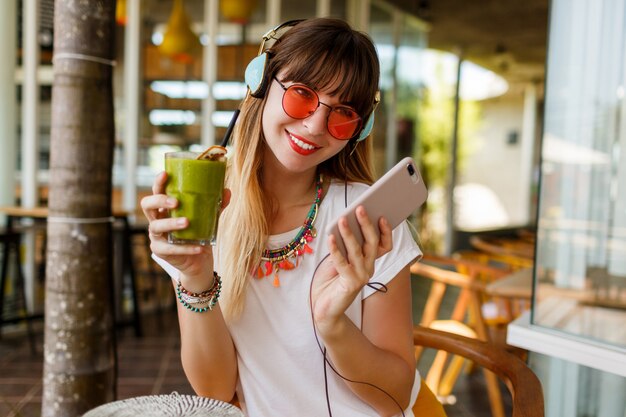 Femme élégante dans des verres roses bénéficiant d'un smoothie sain vert, écoutant de la musique avec des écouteurs, tenant un téléphone mobile.