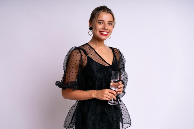 Femme élégante et élégante portant une robe de soirée noire tenant un verre avec une boisson, prête pour la célébration, fond blanc.