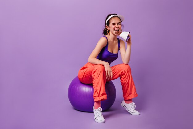 Femme élégante en haut violet et pantalon de survêtement rouge mange une barre de chocolat assis sur fitball contre le mur violet