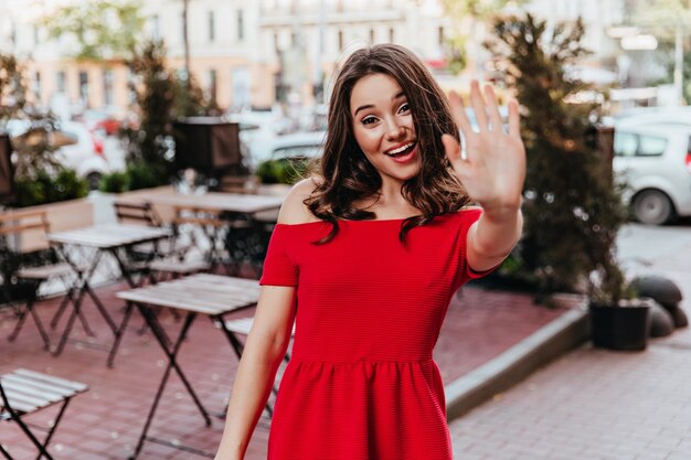 Femme élégante insouciante, agitant la main à la caméra. Portrait en plein air de jolie fille brune en robe rouge debout près du café de la rue.