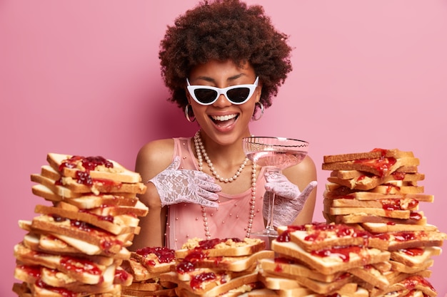 Une femme élégante et joyeuse exprime des émotions positives et s'amuse à la fête, se tient près de la table avec une pile de délicieux toasts de pain, tient un verre de cocktail, porte des lunettes de soleil, une robe et des gants en dentelle