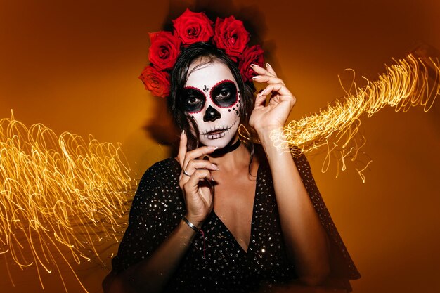 Femme élégante et mince avec des roses dans les cheveux et un masque de crâne posant pour un portrait festif déplaçant ses mains avec élégance