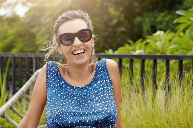 Femme élégante portant des lunettes de soleil et une robe bleue se détendre à l'extérieur contre la forêt verte, ayant un regard heureux et joyeux.