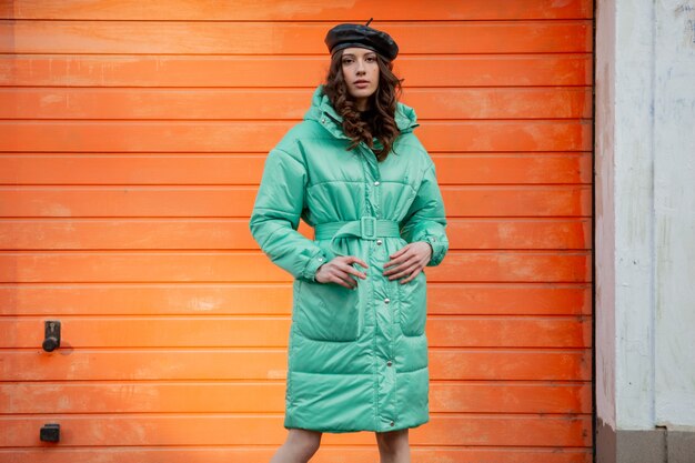 Femme élégante posant en hiver automne mode manteau matelassé et béret chapeau contre le mur orange dans la rue