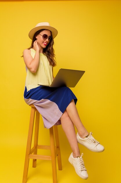 Femme élégante positive en chapeau et robe d'été travaillant avec un ordinateur portable sur un mur jaune