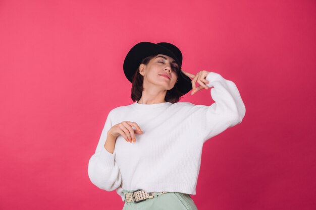 Femme élégante en pull blanc décontracté et chapeau sur un mur rouge rose