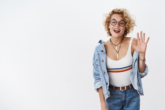 Femme élégante sociable enthousiaste et amicale en lunettes et veste en jean rencontrer des amis agitant joyeusement avec la paume levée en bonjour geste de salutation souriant positif à la caméra sur fond blanc