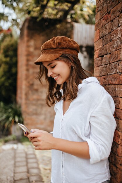 Femme élégante souriante en haut blanc et coiffe brune s'appuie sur le mur de briques et discute au téléphone.