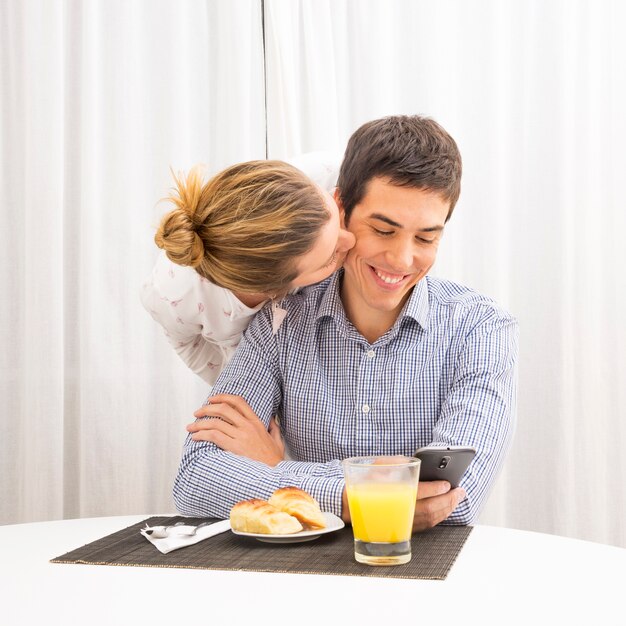 Femme embrasse son mari prenant son petit déjeuner à l&#39;aide d&#39;un téléphone portable