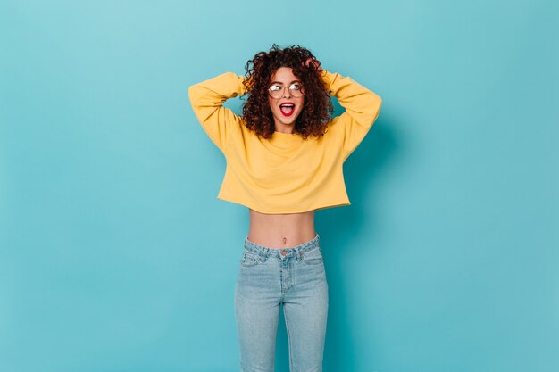 Femme émotionnelle aux cheveux courts et bouclés hurle. Dame en pull jaune et pantalon en jean léger se penche sur la caméra sur l'espace bleu.