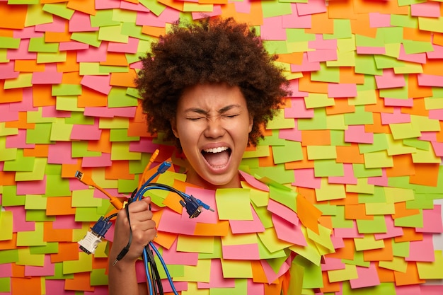 Photo gratuite une femme émotionnelle a besoin de l'aide d'un technicien professionnel, détient de nombreux câbles colorés, ne sait pas comment connecter tous les cordons à l'ordinateur, sort la tête, entourée de nombreux autocollants, a la bouche ouverte