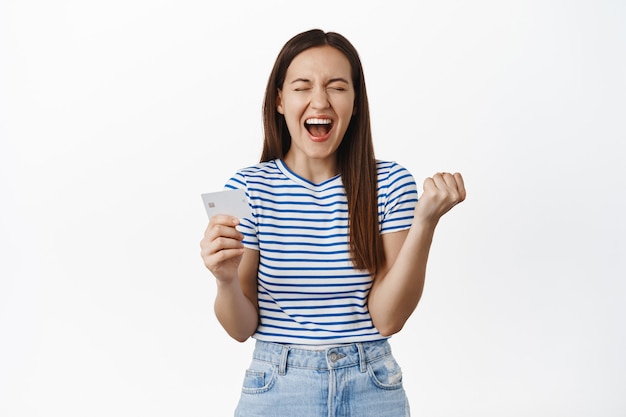 Une femme émotionnelle crie de bonheur et de satisfaction, montrant une carte de crédit, serrant le poing comme un champion, célébrant, debout sur un mur blanc