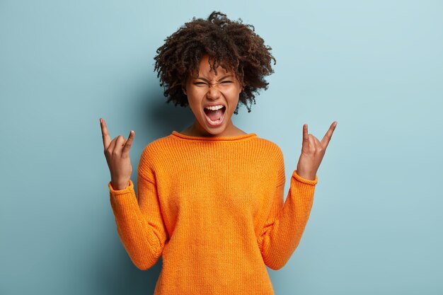 Une femme émotionnelle à la peau sombre fait un geste rock n roll, apprécie la musique cool à la fête, fronce le visage, ouvre la bouche, démontre le geste de la main, vêtue d'un pull orange, modèles sur un mur bleu