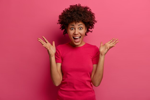 Une femme émotive à la peau sombre a choqué une expression joyeuse, lève les mains, crie fort, soutient l'équipe de football préférée pendant que les montres correspondent, surprise d'entendre d'excellentes nouvelles, porte un t-shirt rose