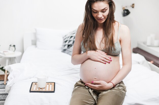 Femme enceinte assise sur le lit en touchant son ventre