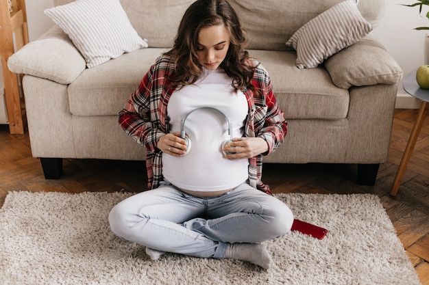 Photo gratuite femme enceinte brune bouclée met des écouteurs sur le ventre