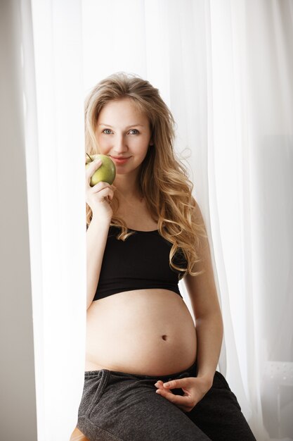 femme enceinte, dans, noir, outfir, séance, à, rebord fenêtre, dans, confortable, chambre à coucher, tenue, ventre, à, main, manger, pomme
